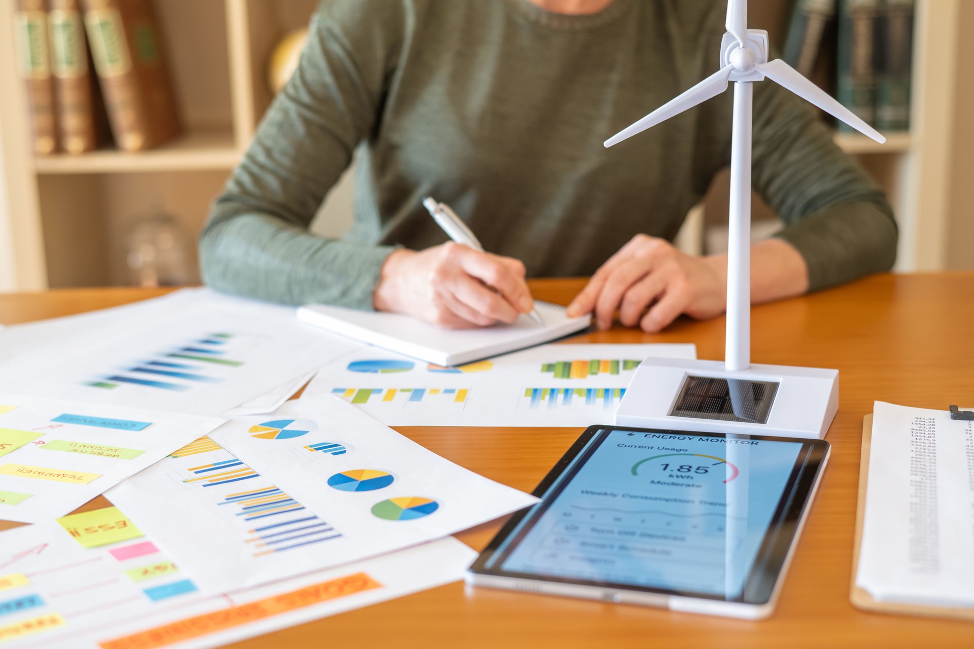 Woman focusing on data analysis and planning for green business and renewable energy, reviewing performance charts on papers and a tablet with a wind turbine model on a wooden office desk